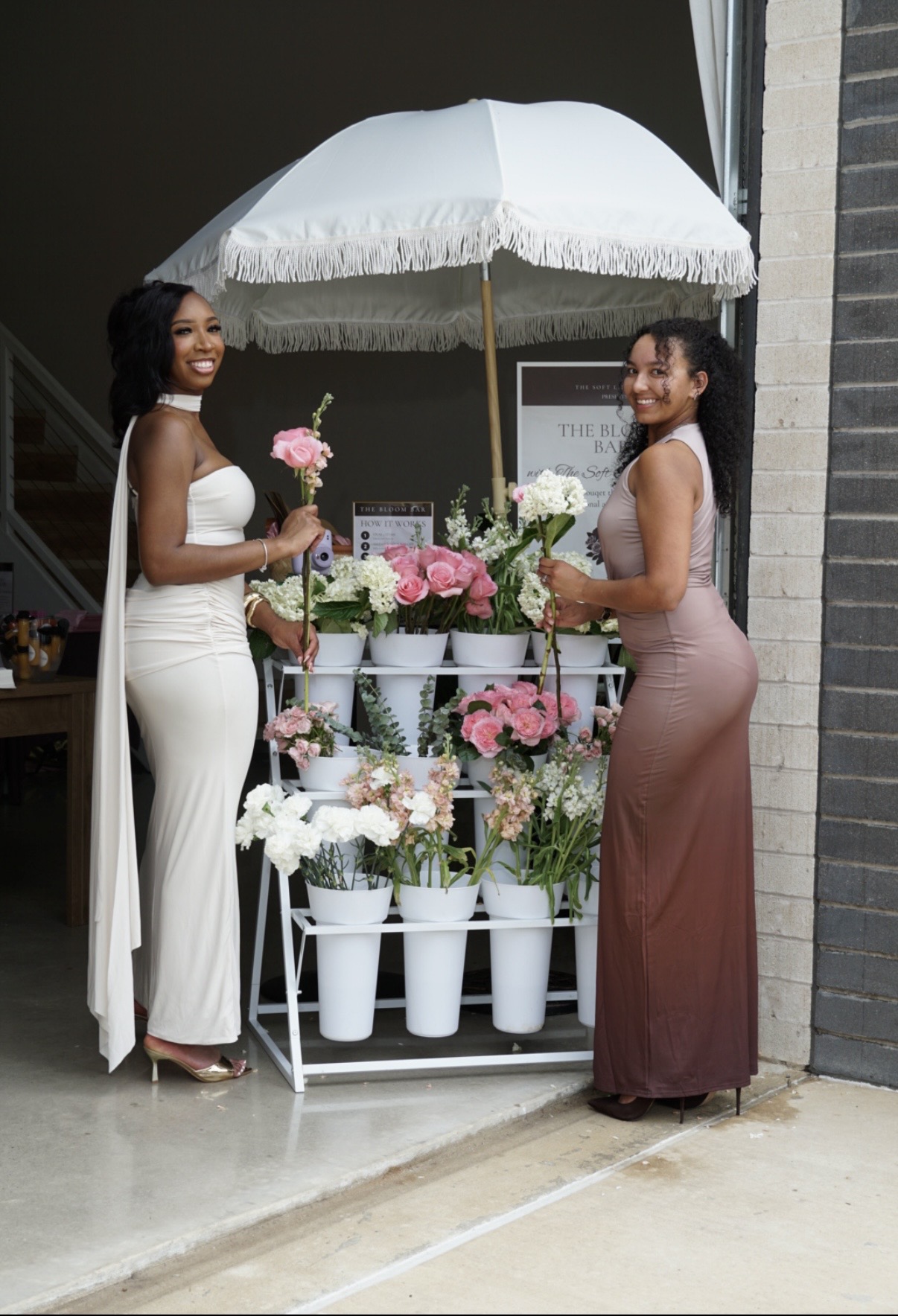 Two women in elegant dresses posing with a floral bloom bar cart under a fringed umbrella