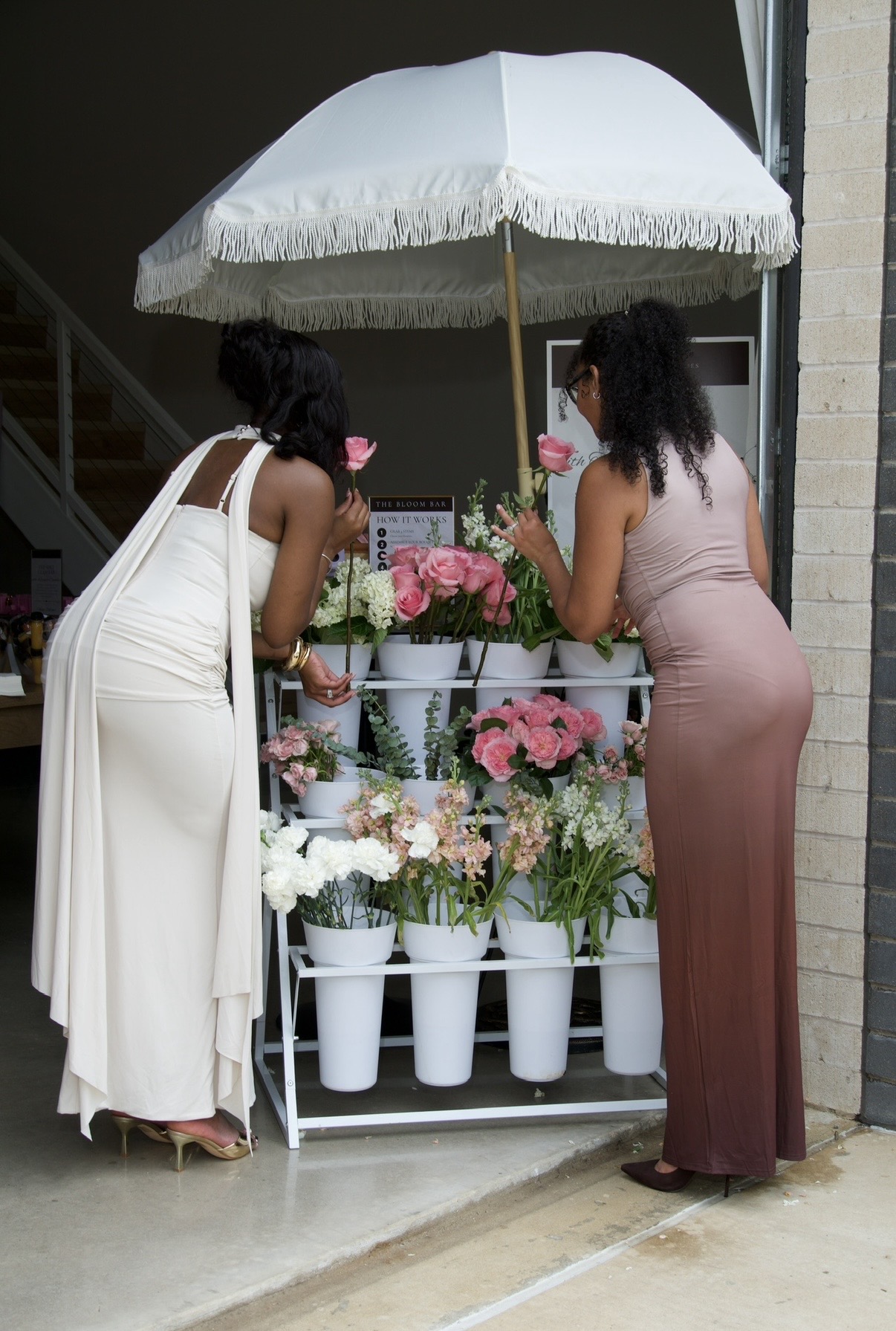 Two women selecting flowers from a bloom bar cart under a fringed umbrella