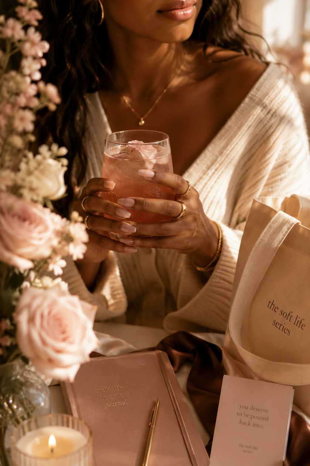 A woman in a cream cardigan holding a rose-topped drink, with a journal, candle, and tote bag nearby